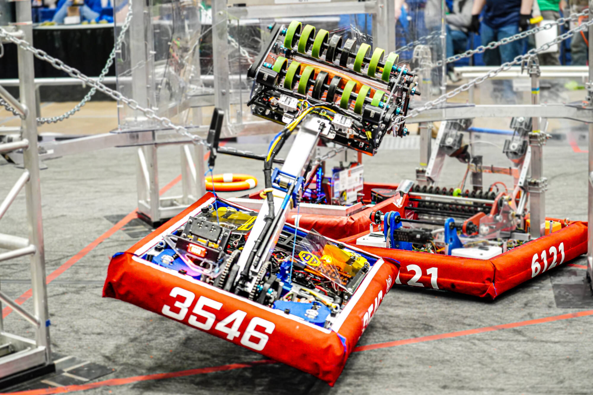 At a GVSU-hosted FIRST Robotics Competition district event, two robots, numbers 3546 and 6121, compete on the field with their lift mechanisms raised.
